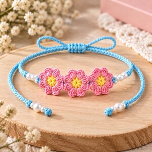 May include: A light blue cord bracelet featuring three crocheted pink flowers with yellow centers. The bracelet is accented with small white beads and includes a decorative blue bow at the top, with adjustable ends finished with white beads.
