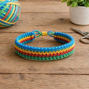 May include: A woven bracelet featuring horizontal stripes of blue, yellow, red, and green yarn. The bracelet has a circular shape with a small yellow knotted closure at the top, showcasing a detailed braided texture throughout the entire piece.
