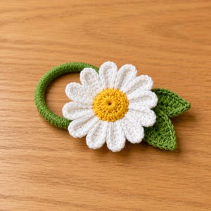 May include: A hair tie featuring a crocheted daisy with white petals and a yellow center, attached to a green circular band. Two small, textured green crocheted leaves are positioned behind the flower, adding detail to the floral design.
