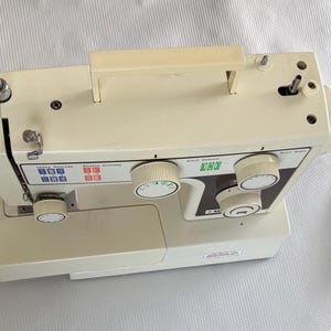 Puede incluir: Una máquina de coser de color crema con un asa de transporte superior, dos portabobinas y un panel de control con tres diales circulares. La máquina incluye un pedal conectado por un cable negro y etiquetas para la selección de puntadas.