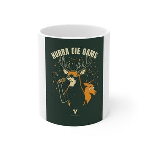 May include: A white ceramic mug featuring a dark green rectangular graphic with the text HURRA DIE GAMS. The illustration depicts a deer wearing orange ski goggles and holding a microphone, accompanied by a smaller deer, surrounded by orange stars.