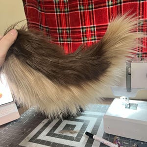 May include: A long, curved faux fur tail featuring a two-tone colour pattern with dark brown fur along the top section and lighter cream-coloured fur along the bottom section, showcasing a soft, shaggy texture throughout the entire length.
