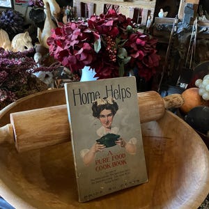 May include: A vintage cookbook titled "Home Helps: A Pure Food Cook Book" with a woman holding a pot on the cover. The book is sitting in a wooden bowl with a rolling pin.
