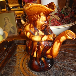May include: A brown ceramic pitcher shaped like a man wearing a hat. The pitcher has a spout shaped like a long, curved horn. The pitcher is sitting on a wooden surface.