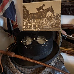 May include: A black top hat with round wire-rimmed glasses, a riding crop, and a vintage photograph of a horse jumping a fence. The photograph is in a frame. A Union Jack flag is in the background.