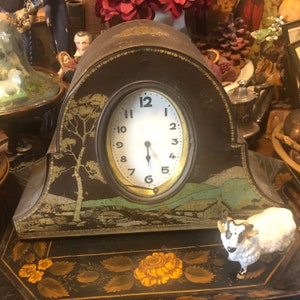 May include: A vintage, brown, metal clock with a white face and black hands. The clock is decorated with a painted landscape scene and a small white sheep figurine. The clock is sitting on a black surface with a floral design.