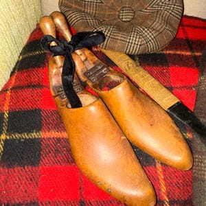 May include: A pair of wooden shoe forms tied with a black ribbon, resting on a red and black plaid blanket. A brown and tan plaid newsboy cap is in the background. A vintage aesthetic.
