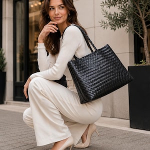 May include: A black tote bag featuring a woven leather construction with a rectangular shape and two shoulder straps. The bag has a consistent grid-like pattern across the entire exterior surface, providing a textured appearance and a structured silhouette.