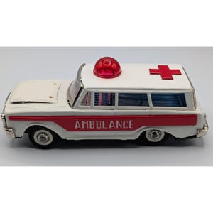 Könnte beinhalten: Ein weißes Spielzeug-Krankenwagenmodell mit einem roten Streifen an der Seite, auf dem das Wort AMBULANCE in weißen Großbuchstaben steht. Das Fahrzeug hat ein rotes Blaulicht auf dem Dach und ein rotes Kreuz auf dem hinteren Teil mit silbernen Rädern.