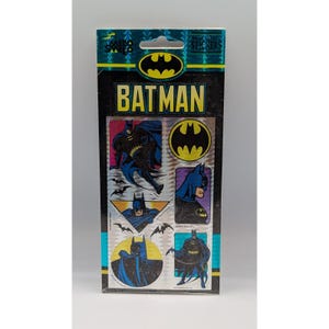 Può includere: Una confezione rettangolare di adesivi rimovibili di Batman con un logo nero e giallo nella parte superiore. La confezione mostra varie illustrazioni di Batman in costumi blu e neri, inclusi disegni circolari, triangolari e rettangolari.