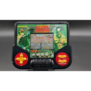 以下が含まれることがあります： Judge Dreddのブランドロゴが入った黒い携帯型電子ゲーム機。中央に液晶画面、赤い円形ベースに黄色い十字キー、Kick、Punch、Pick/Call Fireと書かれた赤いボタンが配置されている。Electronicという文字が記載されている。
