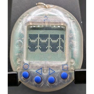 May include: A clear, oval-shaped digital pet device featuring a square LCD screen displaying four pixelated character icons. The handheld electronic toy includes four round, blue control buttons positioned below the screen with blue symbols.