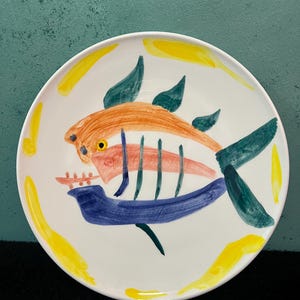 以下が含まれることがあります： 中央に手描きの魚の模様が施された、白い円形の陶磁器製プレート。魚はオレンジ、ダークグリーン、青の筆致で描かれており、プレートの縁は黄色の曲線的なアクセントで装飾されています。
