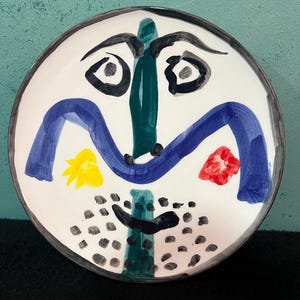 May include: A round ceramic plate featuring a hand-painted abstract face design. The face has a dark green vertical nose, arched blue eyebrows, black eyes, a yellow cheek spot, a red cheek spot, and black dots representing facial hair.