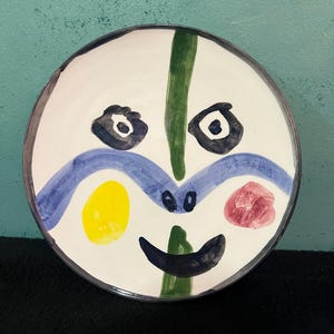 Puede incluir: Un plato de cerámica redondo con base blanca que presenta un diseño de cara abstracta pintado a mano. La cara incluye dos ojos azul oscuro, una línea azul ondulada en el centro, un círculo amarillo a la izquierda, un círculo rosa a la derecha y una sonrisa azul oscuro.