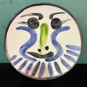 Op de afbeelding: Een ronde keramische bord met een witte achtergrond en een handgeschilderd abstract gezicht. Het ontwerp bevat twee zwarte cirkelvormige ogen met witte kruisdetails, een centrale groene neus, blauwe gebogen lijnen en een donkerblauw kraagachtig patroon.