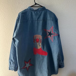 May include: A light blue denim button-down shirt featuring a large central applique of a cowboy boot with a red foot and a red and white checkered shaft. Two five-pointed star appliques are stitched onto the back, one red and one grey.