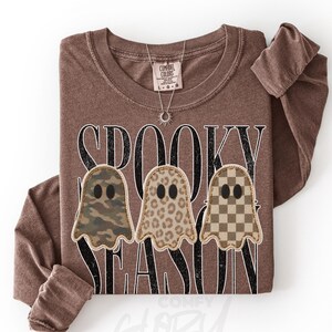 Può includere: Una maglietta a maniche lunghe marrone con la scritta SPOOKY SEASON in lettere nere. Sotto il testo ci sono tre illustrazioni di fantasmi con motivi diversi: uno mimetico, uno leopardato e uno a scacchi marroni e beige.