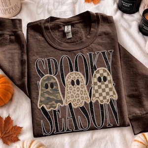 May include: A dark brown crewneck sweatshirt featuring the text SPOOKY SEASON in large white letters. Three ghost illustrations are centred on the front, each with a different pattern: camouflage, leopard print, and a tan and white checkerboard.