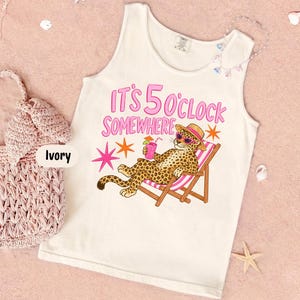 Puede incluir: Una camiseta de tirantes color marfil con un gráfico de un leopardo con sombrero y gafas de sol, descansando en una silla de playa con una bebida. El texto dice IT'S 5 O'CLOCK SOMEWHERE. El diseño incluye estrellas rosas y naranjas, y se muestra junto a un bolso de ganchillo rosa.