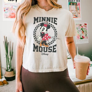 May include: An ivory boxy t-shirt featuring a graphic of Minnie Mouse inside a black and white checkered circle. The text reads MINNIE MOUSE and Disney, with a small Minnie Mouse 2020 signature. The shirt is labeled Comfort Colors Boxy Tee.