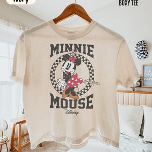 May include: An ivory boxy t-shirt featuring a graphic of Minnie Mouse inside a black and white checkered circle. The text reads MINNIE MOUSE and Disney. The shirt is labeled Comfort Colors Boxy Tee and Ivory. It has short sleeves and a crew neck.