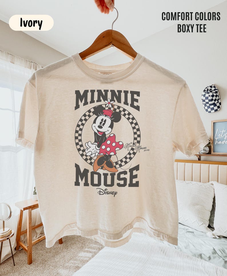 May include: An ivory boxy t-shirt featuring a graphic of Minnie Mouse inside a black and white checkered circle. The text reads MINNIE MOUSE and Disney. The shirt is labeled Comfort Colors Boxy Tee and Ivory. It has short sleeves and a crew neck.