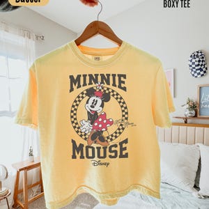 May include: A butter yellow boxy t-shirt featuring a graphic of Minnie Mouse inside a black and yellow checkered circle. The text reads MINNIE MOUSE and Disney. The shirt is displayed on a wooden hanger against a light-colored bedroom background.