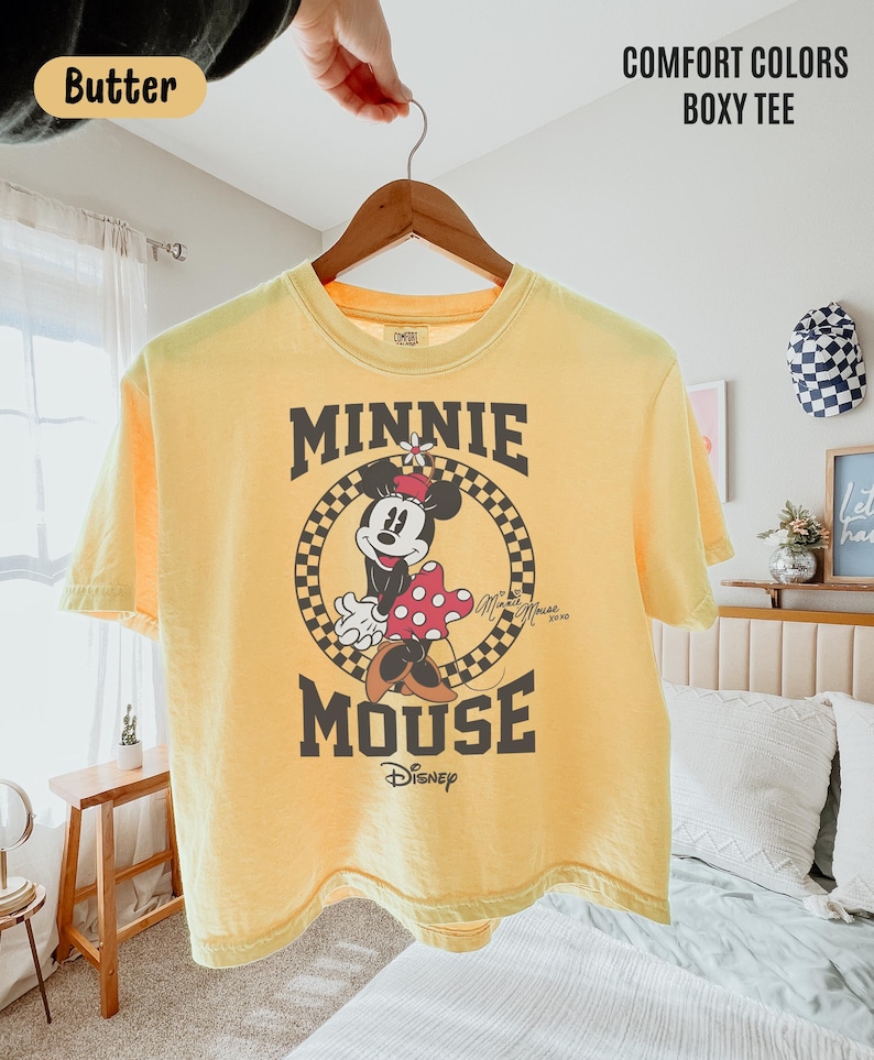 May include: A butter yellow boxy t-shirt featuring a graphic of Minnie Mouse inside a black and yellow checkered circle. The text reads MINNIE MOUSE and Disney. The shirt is displayed on a wooden hanger against a light-colored bedroom background.