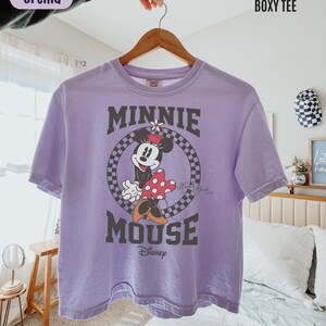 May include: An orchid-colored short-sleeve boxy t-shirt featuring a graphic of Minnie Mouse inside a black and white checkered circle. The text "MINNIE MOUSE" is printed in large dark letters above and below the graphic, with "Disney" below.