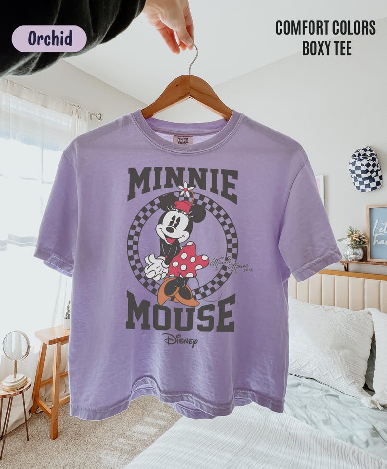 May include: An orchid-colored short-sleeve boxy t-shirt featuring a graphic of Minnie Mouse inside a black and white checkered circle. The text "MINNIE MOUSE" is printed in large dark letters above and below the graphic, with "Disney" below.