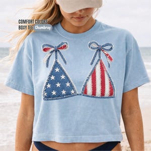 May include: A light blue boxy t-shirt featuring a graphic of a bikini top with a patriotic American flag design. The left cup displays white stars on a blue background, while the right cup features red and white vertical stripes. Text reads COMFORT COLORS BOXY TEE Chambray.