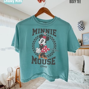 May include: A chalky mint boxy t-shirt featuring a graphic of Minnie Mouse inside a black and white checkered circle. The text Minnie Mouse and Disney are printed in dark gray. The shirt has a crew neckline and short sleeves on a hanger.