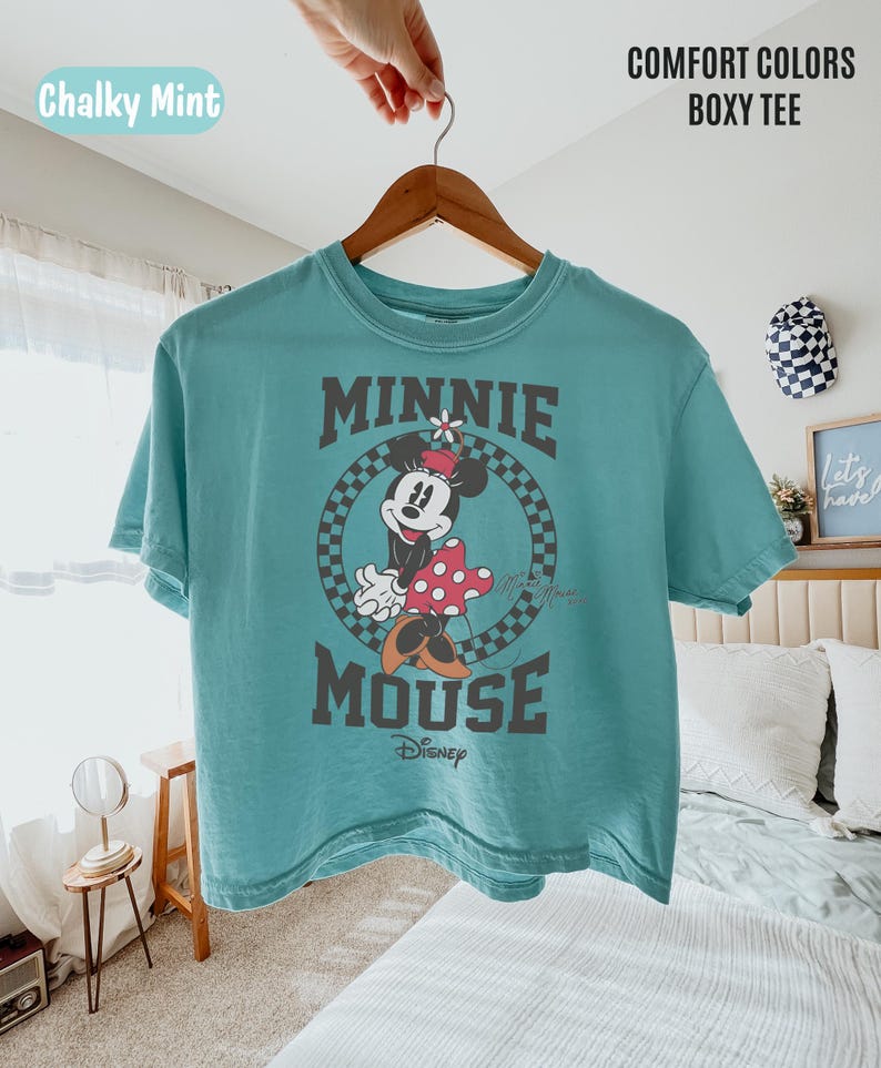 May include: A chalky mint boxy t-shirt featuring a graphic of Minnie Mouse inside a black and white checkered circle. The text Minnie Mouse and Disney are printed in dark gray. The shirt has a crew neckline and short sleeves on a hanger.