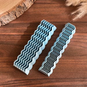 Puede incluir: Dos herramientas rectangulares de color azul claro con una textura de superficie ondulada y acanalada. Los objetos son alargados y presentan un patrón repetitivo de crestas paralelas a lo largo de toda su longitud, con una base plana y un borde superior texturizado.