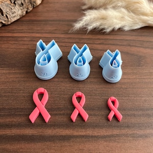 May include: Three light blue plastic ribbon-shaped cookie cutters in sizes 30, 25, and 20, accompanied by three corresponding pink ribbon-shaped fondant or dough cutouts in descending sizes, arranged in two rows on a dark wood surface.