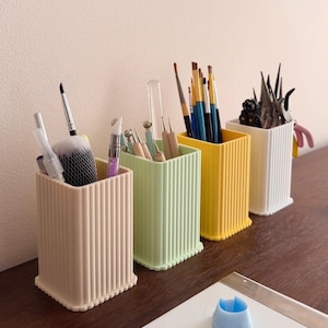 Peut inclure: Quatre organisateurs de bureau rectangulaires à côtes verticales, de couleurs beige pastel, vert clair, jaune et blanc. Chaque récipient contient divers outils de loisirs créatifs, dont des pinceaux, des stylets et des pinces.