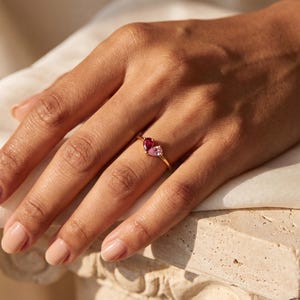May include: A gold ring featuring two pear-cut gemstones, one deep red and one light pink, set side-by-side on a thin, polished gold band. The stones are arranged in a delicate, asymmetrical design that sits horizontally across the finger.