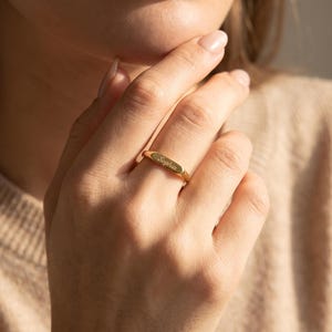 Könnte beinhalten: Ein goldfarbener Metallring mit einem flachen, rechteckigen Mittelstück. Die Oberfläche des Stegs ist mit dem Namen Sophia in einer kursiven Schriftart graviert. Der Ring ist schmal und poliert und weist ein einheitliches Metallfinish auf.