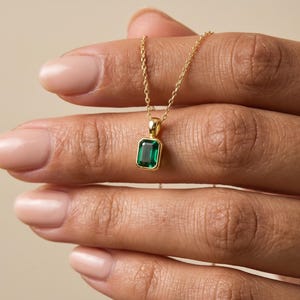 May include: A gold necklace featuring a rectangular emerald-cut green gemstone pendant set in a polished gold bezel. The pendant hangs from a delicate gold chain, showcasing a vibrant green stone with a clear, faceted surface and gold metal.