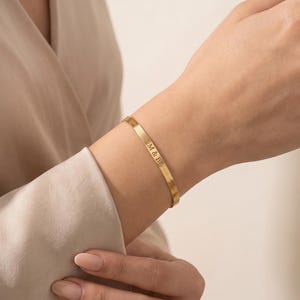 May include: A thin, gold-toned metal cuff bracelet featuring a smooth, polished finish. The bracelet is engraved with the letters M & B in a simple, sans-serif font, positioned vertically along the outer surface of the band.