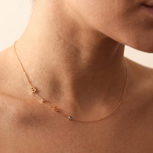 May include: A delicate gold-tone chain necklace featuring a small letter B charm, a tiny pink gemstone, a letter S charm, and a small blue gemstone arranged asymmetrically along the thin metal link chain resting against the collarbone area.