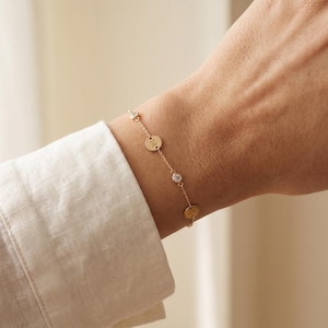 May include: A delicate gold chain bracelet featuring alternating small circular charms and clear round stones. One of the circular charms is engraved with the letter M. The bracelet is secured with a thin gold chain and a clasp mechanism.