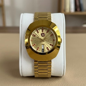 May include: A gold-tone Rado DiaStar wristwatch featuring a sunburst gold dial with red stone hour markers, a day-date window at the six o'clock position, and a matching gold-tone metal link bracelet with a polished, oval-shaped watch case.
