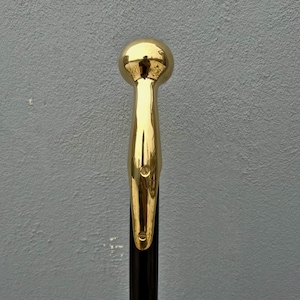 May include: A walking stick featuring a polished gold-tone metal handle with a spherical top and a smooth, elongated neck that tapers downward. The handle is securely attached to a dark, cylindrical shaft, showcasing a reflective, metallic finish.