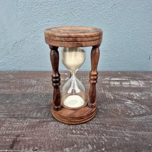 Könnte beinhalten: Eine Sanduhr mit einem dunkelbraunen Holzrahmen, bestehend aus einem runden Ober- und Unterteil, die durch vier gedrechselte Säulen verbunden sind. Ein klarer Glaskolben ist mittig platziert und enthält feinen, cremefarbenen Sand.