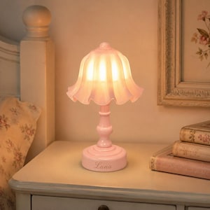 Peut inclure: Une lampe de table rose avec un abat-jour cannelé en forme de cloche diffusant une lueur chaude et douce. La lampe présente un pied rose fin assorti et un socle circulaire avec le prénom Luna gravé en écriture cursive sur la face avant.