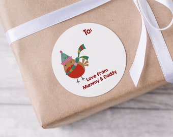 Pode incluir: Uma etiqueta de presente redonda branca com um pintarroxo vermelho usando um chapéu e uma echarpe verdes, azuis e brancos. A etiqueta diz "Para:" e "Com amor de Mamãe e Papai".