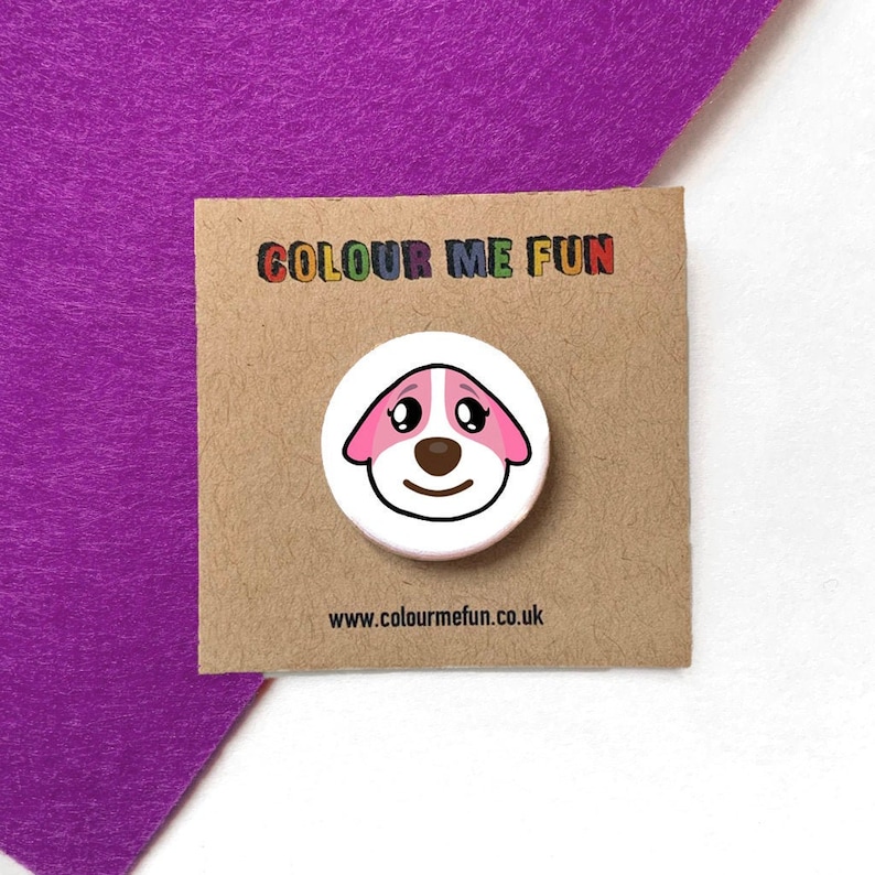 Puede incluir: Un bot&oacute;n blanco con la cara de un perro de dibujos animados, orejas y detalles rosas, una nariz marr&oacute;n y una boca sonriente. El bot&oacute;n est&aacute; sobre una tarjeta marr&oacute;n con el texto "COLOUR ME FUN" y "www.colourmefun.co.uk". El fondo es morado.