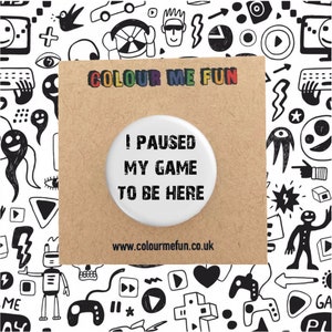 Könnte beinhalten: Weißer Knopf mit schwarzem Text, der "Ich habe mein Spiel pausiert, um hier zu sein" lautet. Der Knopf befindet sich auf einer braunen Karte mit dem Text "COLOUR ME FUN" in Regenbogenfarben und der Website-Adresse "www.colourmefun.co.uk".